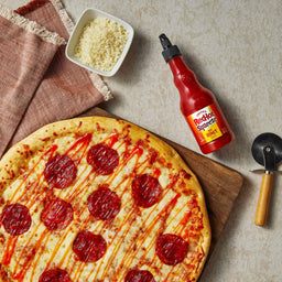 ["A pepperoni pizza with Frank's RedHot Squeeze Hot Honey Sauce on a wooden cutting board with a pizza cutter and a bowl of shredded cheese."]