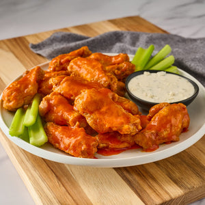 A white plate holds a generous serving of chicken wings coated in a vibrant orange sauce made with Franks Rdhot Bws Gb 24 148 Ml alongside fresh celery sticks and a bowl of creamy blue cheese dressing.