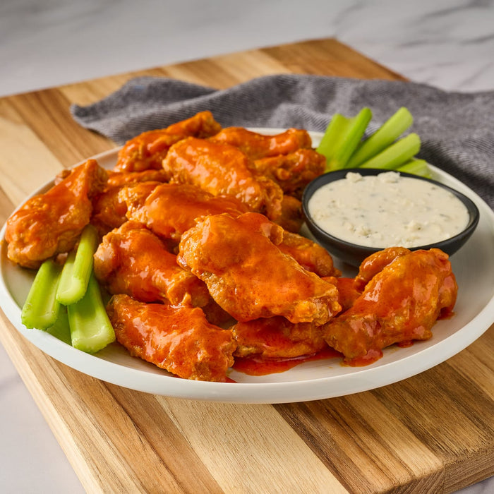 A white plate holds a generous serving of chicken wings coated in a vibrant orange sauce made with Franks Rdhot Bws Gb 24 148 Ml alongside fresh celery sticks and a bowl of creamy blue cheese dressing.