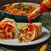 A stream of Cholula Original Mexican Hot Sauce 150Ml is poured from its iconic bottle onto a hearty chicken burrito filled with avocado and cheese, served alongside a bowl of seasoned Mexican rice.