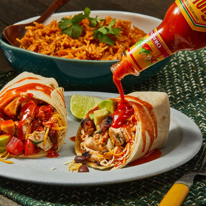 A stream of Cholula Original Mexican Hot Sauce 150Ml is poured from its iconic bottle onto a hearty chicken burrito filled with avocado and cheese, served alongside a bowl of seasoned Mexican rice.