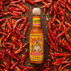 A bottle of Cholula Original Mexican Hot Sauce 150Ml with its signature wooden cap is centered on a dark wooden surface surrounded by a vibrant scattering of dried red chili peppers and seeds.