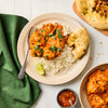 A plate of creamy orange chicken curry sits on a bed of white rice, garnished with cilantro and served with naan bread and a lime wedge on a white wooden table.
