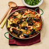 A large paella pan is filled with a colorful rice dish containing chicken, shrimp, mussels, and peas, served with lemon wedges and a side salad.