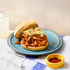 A pulled pork sandwich topped with coleslaw sits on a blue plate, with a small yellow bowl of paprika and a glass of lemonade nearby.