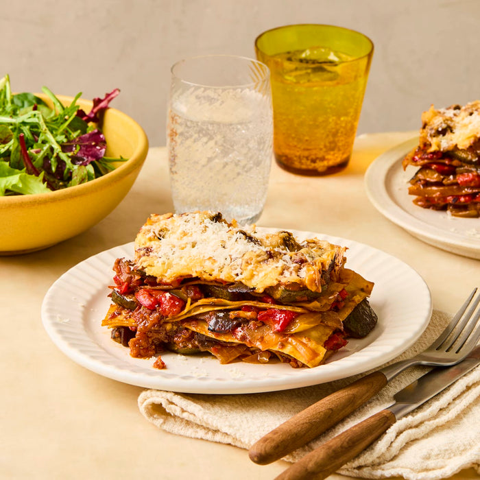 A slice of vegetable lasagna sits on a white plate, served with a side salad and a glass of sparkling water.