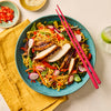 A vibrant chicken noodle stir-fry with sliced grilled chicken and fresh vegetables is served in a blue bowl with pink chopsticks.
