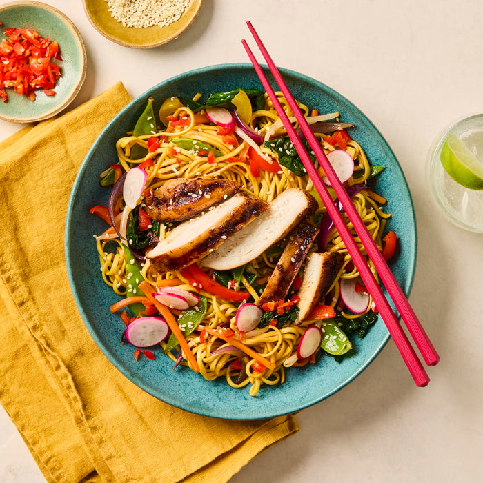 A vibrant chicken noodle stir-fry with sliced grilled chicken and fresh vegetables is served in a blue bowl with pink chopsticks.