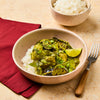 A bowl of green curry with chicken, snow peas, and eggplant is served over white rice, with a lime wedge for garnish.