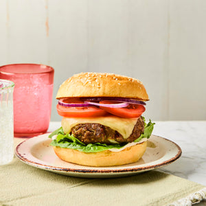 A juicy cheeseburger with lettuce, tomato, and onion on a sesame seed bun is served on a plate with drinks in the background.