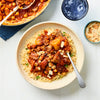 A rustic bowl of couscous is topped with a rich lamb and chickpea stew, garnished with sliced almonds, and served on a white wooden table.