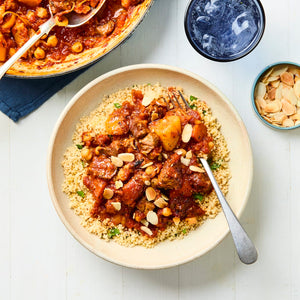 A rustic bowl of couscous is topped with a rich lamb and chickpea stew, garnished with sliced almonds, and served on a white wooden table.