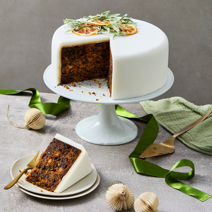 A festive Christmas fruitcake covered in white fondant is displayed on a cake stand with a slice cut out and served on a plate, surrounded by green ribbon and holiday ornaments.