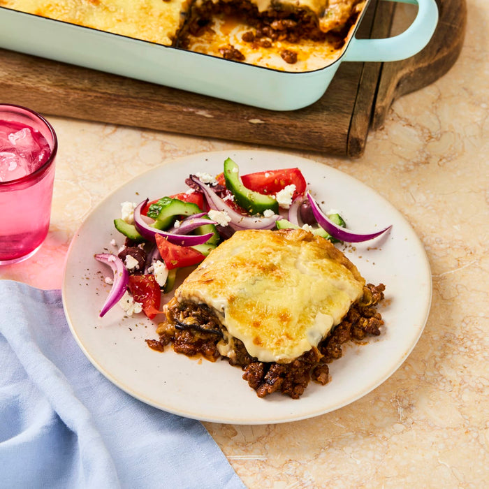 A serving of golden-brown baked moussaka is presented on a white plate next to a fresh Greek salad and a pink iced beverage.