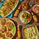 Mexican food platter with tacos, rice, and a green dish on a wooden table with hot sauce bottles.