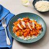 A high-angle shot of a teal bowl filled with white rice, a colorful vegetable stew, and topped with slices of seared chicken breast, with a blue napkin and fork to the side.