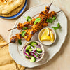 Two skewers of grilled chicken tikka are served on a platter with a side of naan bread, a cucumber and onion salad, and lemon wedges.