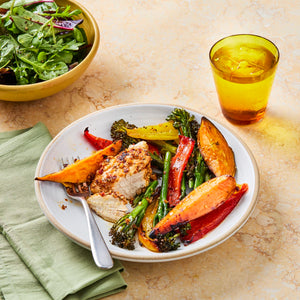 A crusted chicken breast is sliced on a plate and served with roasted sweet potatoes, bell peppers, and broccolini, with a side salad and a drink in the background.