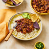 A yellow bowl filled with white rice is topped with a hearty portion of chili con carne, guacamole, and tortilla chips, with a fork resting in the dish.