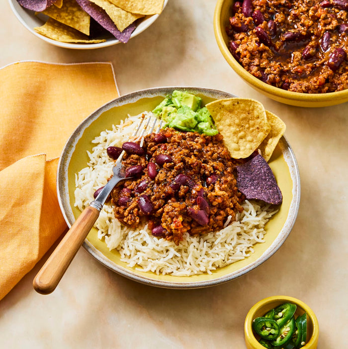 A yellow bowl filled with white rice is topped with a hearty portion of chili con carne, guacamole, and tortilla chips, with a fork resting in the dish.