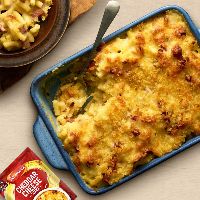 A packet of Schwartz Cheddar Cheese Sauce 38G sits in front of a delicious, golden-brown baked macaroni and cheese with bacon, showing a perfect meal idea for the product.