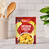 A packet of Schwartz Cheddar Cheese Sauce 38G sits on a white marble kitchen counter, with a utensil holder and a green plant blurred in the background.