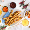 A festive overhead shot shows golden-brown cheese straws, made with Schwartz Cheddar Cheese Sauce 38G, piled on a platter next to two dipping sauces and drinks, with Christmas decorations scattered around.