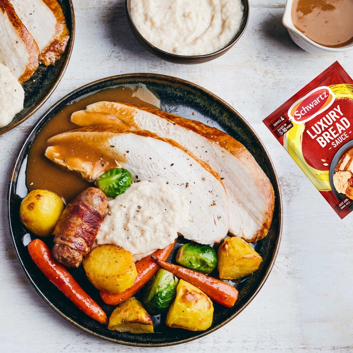 A packet of Schwartz Luxury Bread Sauce 40G is shown next to a plated roast turkey dinner, which includes the prepared sauce, demonstrating a perfect serving suggestion.