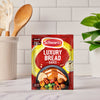 A packet of Schwartz Luxury Bread Sauce 40G sits on a kitchen countertop, with a utensil holder and a small plant blurred in the background.