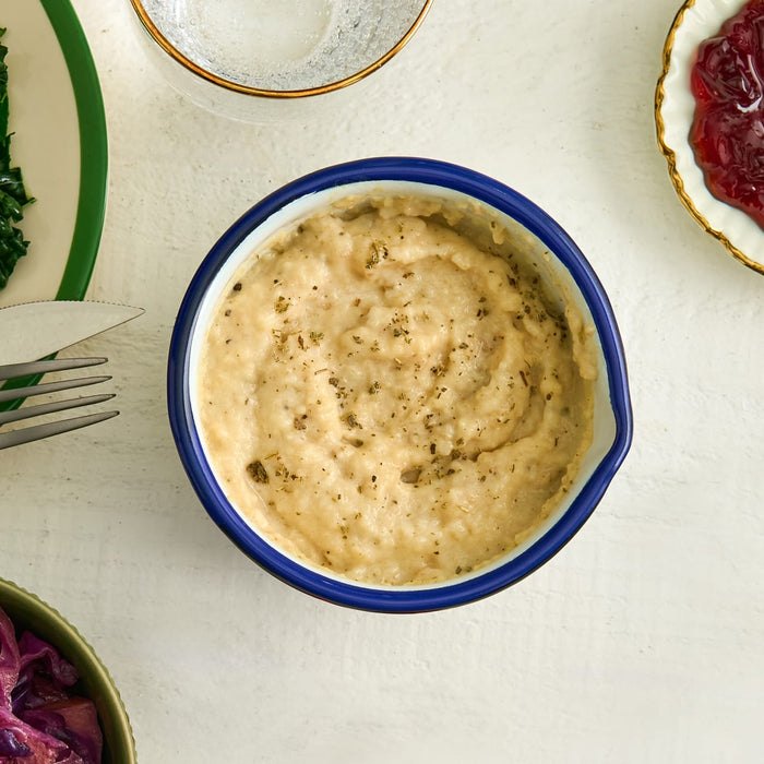 A blue-rimmed bowl filled with prepared Schwartz Luxury Bread Sauce 40G sits on a white table, surrounded by other dishes for a meal.