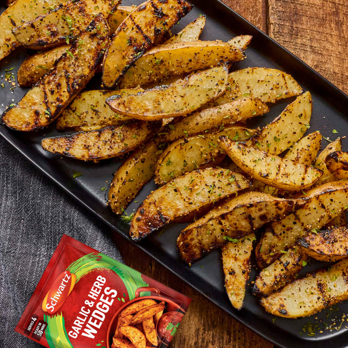 A black platter filled with grilled and seasoned potato wedges serves as a serving suggestion for the accompanying packet of Schwartz Garlic And Herb Wedges, 38G.
