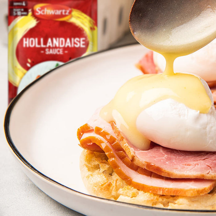 A creamy hollandaise sauce is poured from a spoon over a poached egg and ham on an English muffin, with a packet of Schwartz Hollandaise Sauce Mix 25G visible in the background.