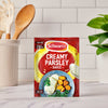 A packet of Schwartz Creamy Parsley Sauce 26G is centered on a kitchen countertop, with the packaging featuring a serving suggestion of fish topped with the creamy sauce.