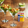 Two martini glasses filled with seasoned shrimp and lettuce sit on a wooden board next to a tin of Old Bay Seasoning and a bowl of lime wedges.