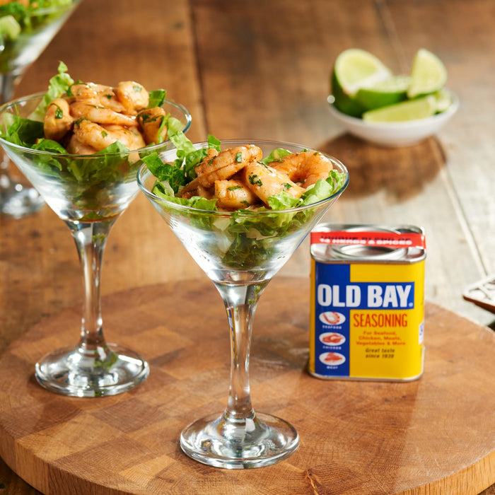 Two martini glasses filled with seasoned shrimp and lettuce sit on a wooden board next to a tin of Old Bay Seasoning and a bowl of lime wedges.