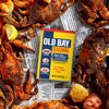 A yellow and blue tin of Old Bay Seasoning with 30 percent less sodium is centered among steamed crabs and potatoes seasoned with spices on a newspaper background.