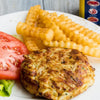 A golden brown crab cake seasoned with Old Bay is served on a plate with crinkle cut fries and a fresh tomato slice.