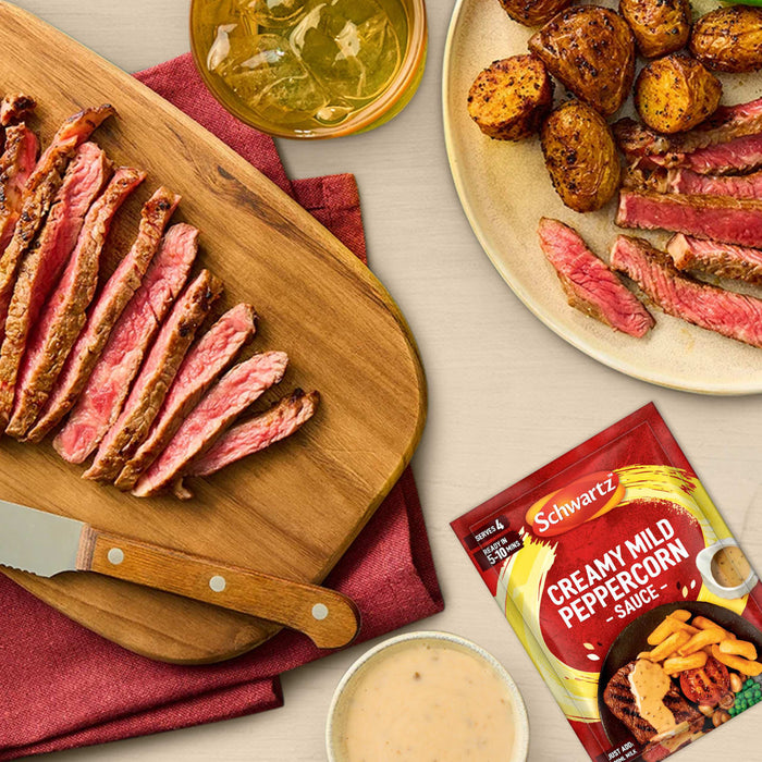 A delicious meal of sliced steak and roasted potatoes is served alongside a packet of Schwartz Creamy Mild Peppercorn Sauce Mix 25G, which can be used to create the accompanying creamy sauce.