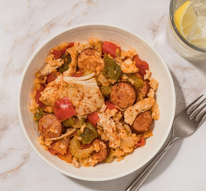 A bowl of chicken and sausage jambalaya seasoned with Schwartz Extra Spicy Cajun 42G Grill&Sizzl sits on a marble surface next to a glass of lemon water.