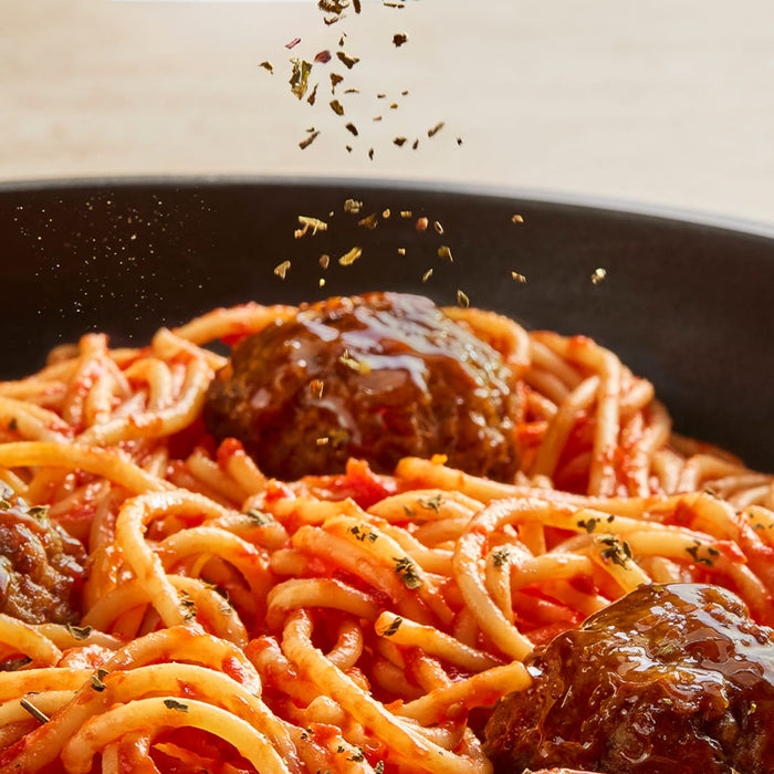 A generous sprinkle of Schwartz Oregano 7G is added as a finishing touch to a close-up shot of a pan filled with spaghetti and meatballs.