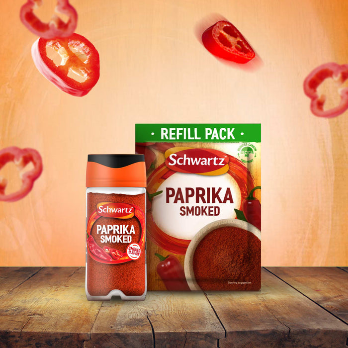A jar of Schwartz Smoked Paprika 40G and its corresponding refill pack sit on a rustic wooden table, with slices of red pepper floating in the background.