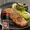 A seasoned salmon fillet served with charred bok choy on a dark plate alongside a bottle of Schwartz Black Peppercorns Adjustable Medium Grinder 35G.