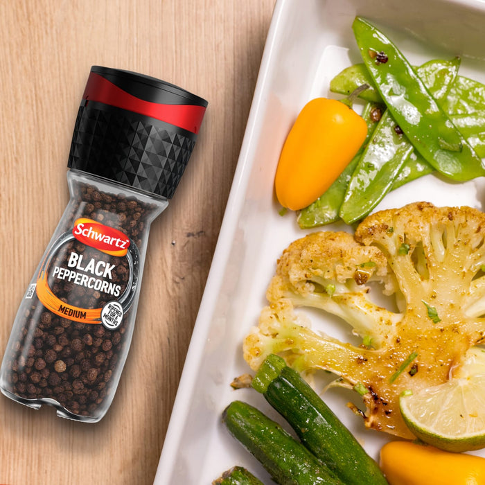 A bottle of Schwartz Black Peppercorns Adjustable Medium Grinder 35G sits on a wooden surface next to a white dish filled with roasted cauliflower, zucchini, and peppers.