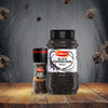 A Schwartz Black Peppercorns Adjustable Medium Grinder 35G stands next to a large catering-sized jar of whole black peppercorns on a rustic wooden surface with peppercorns floating in the air against a dark background.