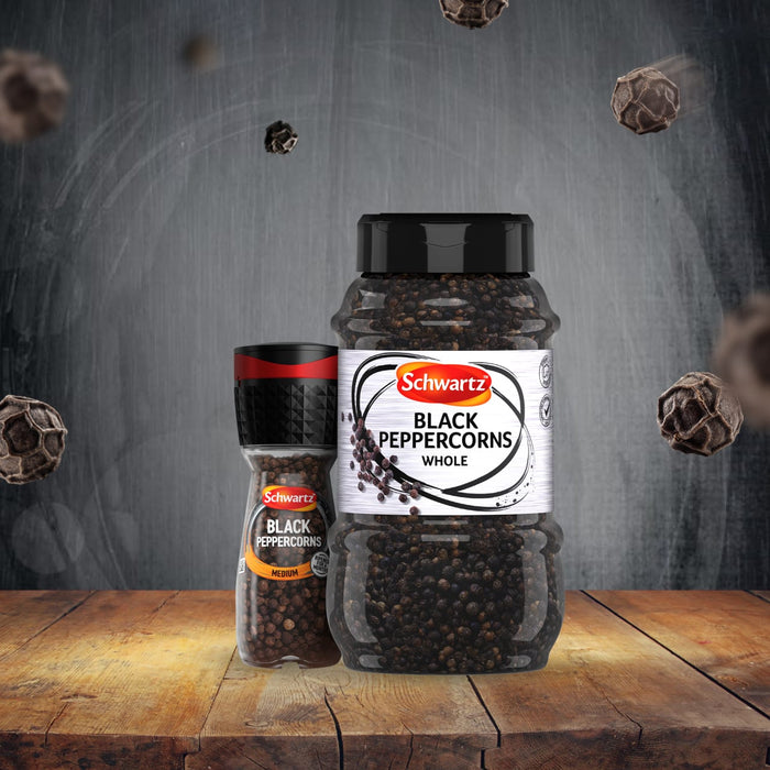 A Schwartz Black Peppercorns Adjustable Medium Grinder 35G stands next to a large catering-sized jar of whole black peppercorns on a rustic wooden surface with peppercorns floating in the air against a dark background.