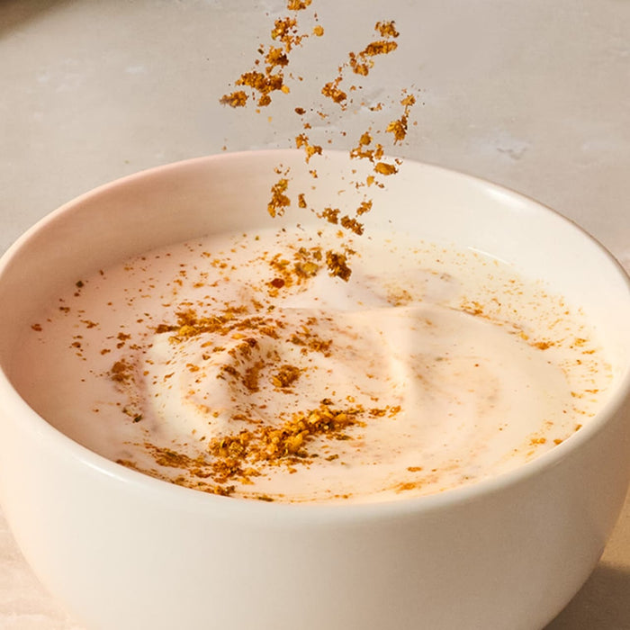 A spoonful of Schwartz Turmeric Ground 37G is being sprinkled over a creamy white dip in a white bowl.