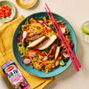 A vibrant bowl of stir-fried noodles and vegetables topped with sliced chicken seasoned with Schwartz Af Chinese Cs Sea sits next to a glass of lime water and small bowls of chili and sesame seeds.