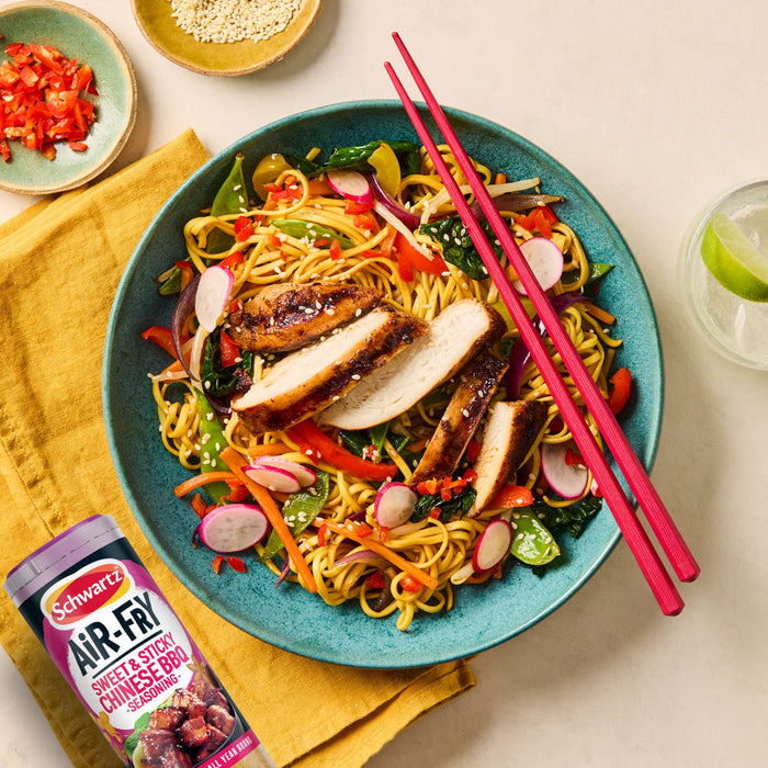 A vibrant bowl of stir-fried noodles and vegetables topped with sliced chicken seasoned with Schwartz Af Chinese Cs Sea sits next to a glass of lime water and small bowls of chili and sesame seeds.