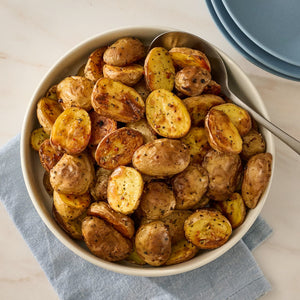 A white serving bowl filled with seasoned and golden brown Jr Ps Roast Potato X6 sits on a blue napkin next to a stack of plates.