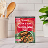 A packet of the Sa Rt Chicken Satay X12 seasoning mix from Schwartz is displayed on a kitchen counter next to a finished dish of chicken satay skewers and peanut sauce.